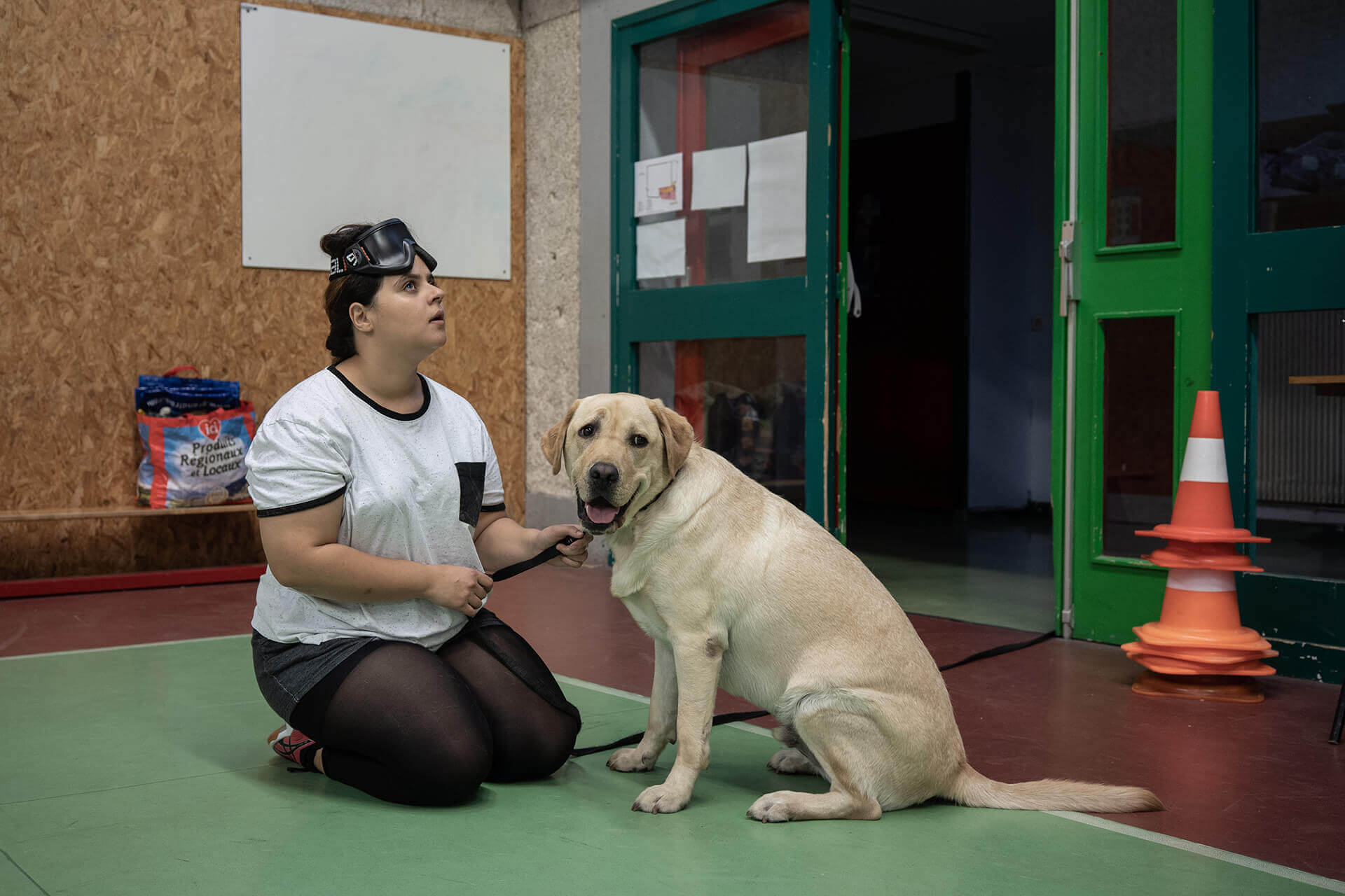 Meryem, jeune-femme non-voyante, est assise face à Mystère, son chien guide. Son visage est tendu vers le haut tandis que Mystère regarde à droite de la photo. Derrière eux, l’entrée du gymnase.