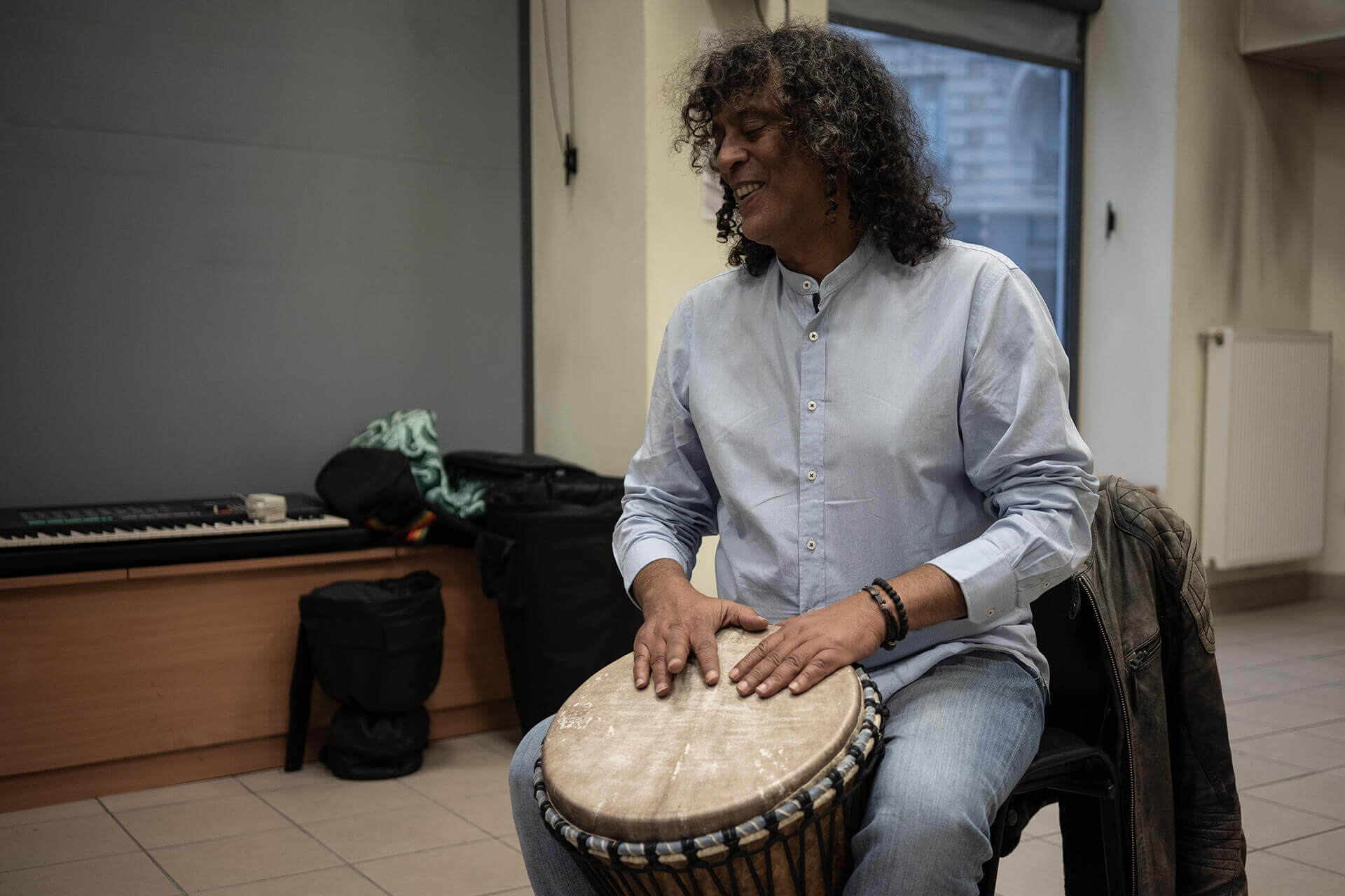 Khaled, professeur de djembé non-voyant, est assis sur une chaise en train de jouer de son instrument. Il sourit, la tête légèrement inclinée vers sa droite. Derrière lui, un piano électrique repose dans un coin de la salle.